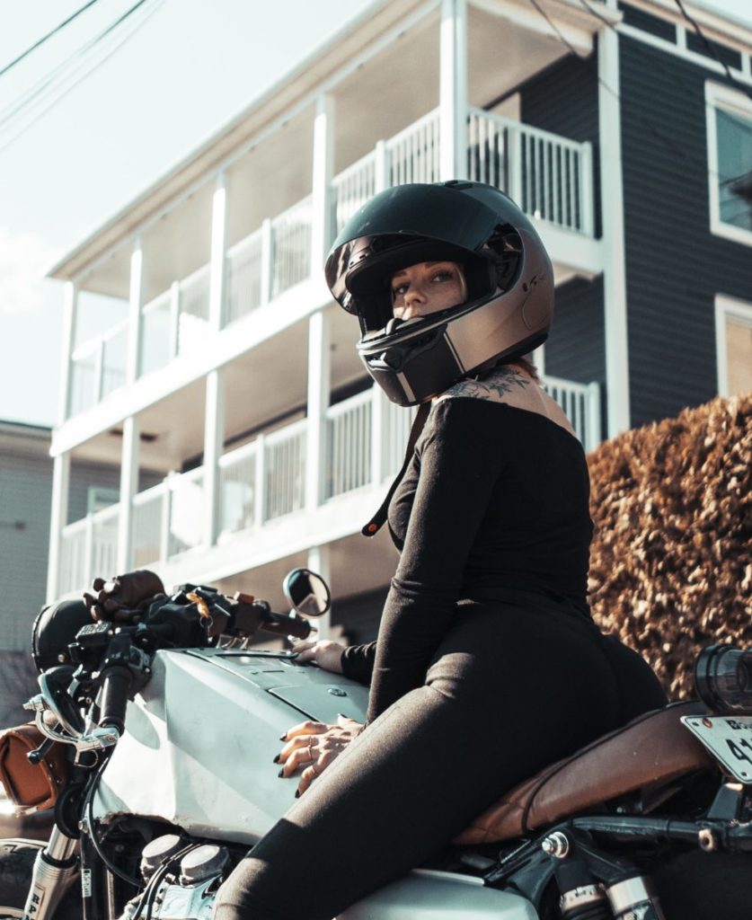 Woman in black clothes and a gold helmet on a light green motorcycle stopped in front of a large house