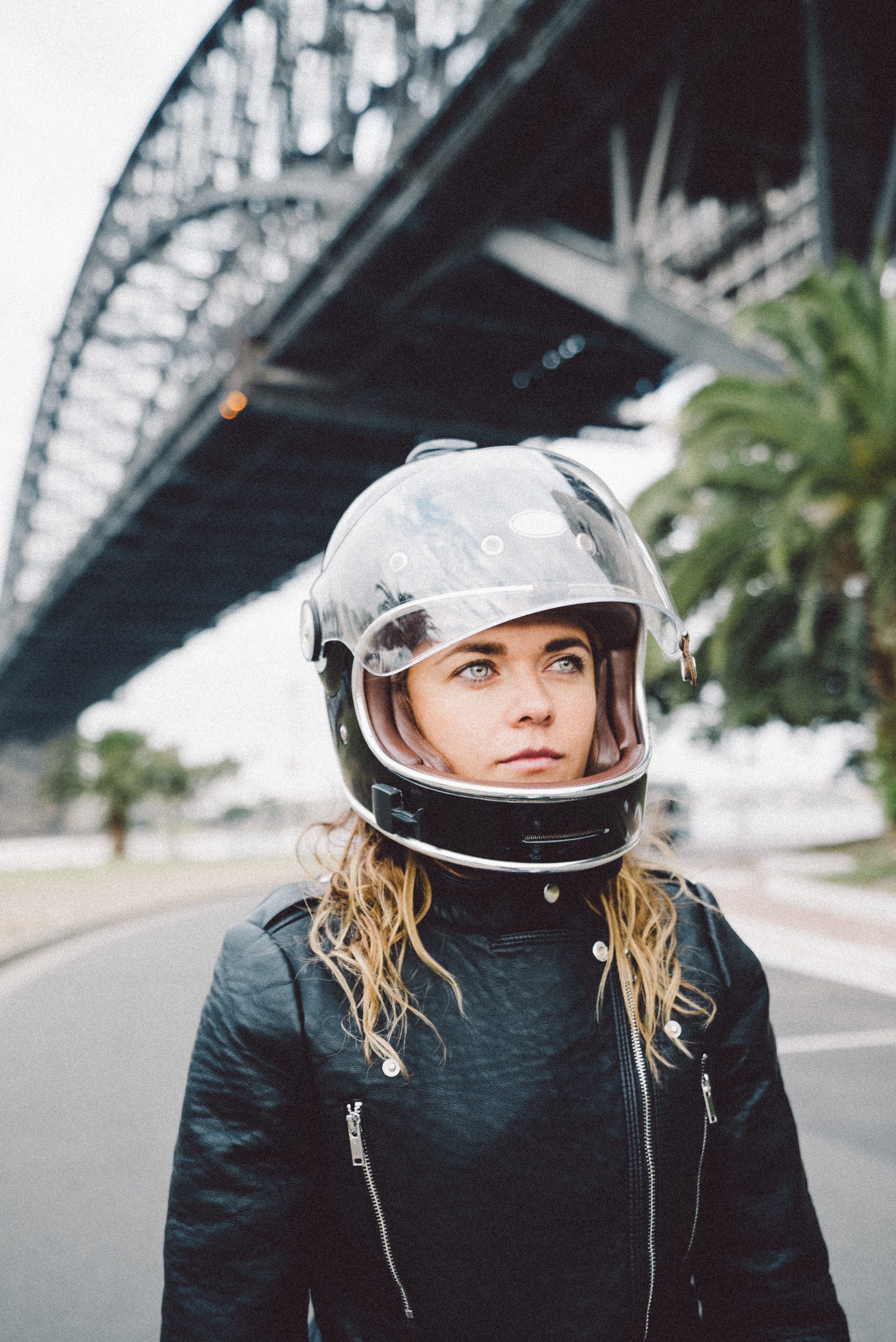 Blond woman in front of a bridge in a black leather jacket and black motorcycle helmet