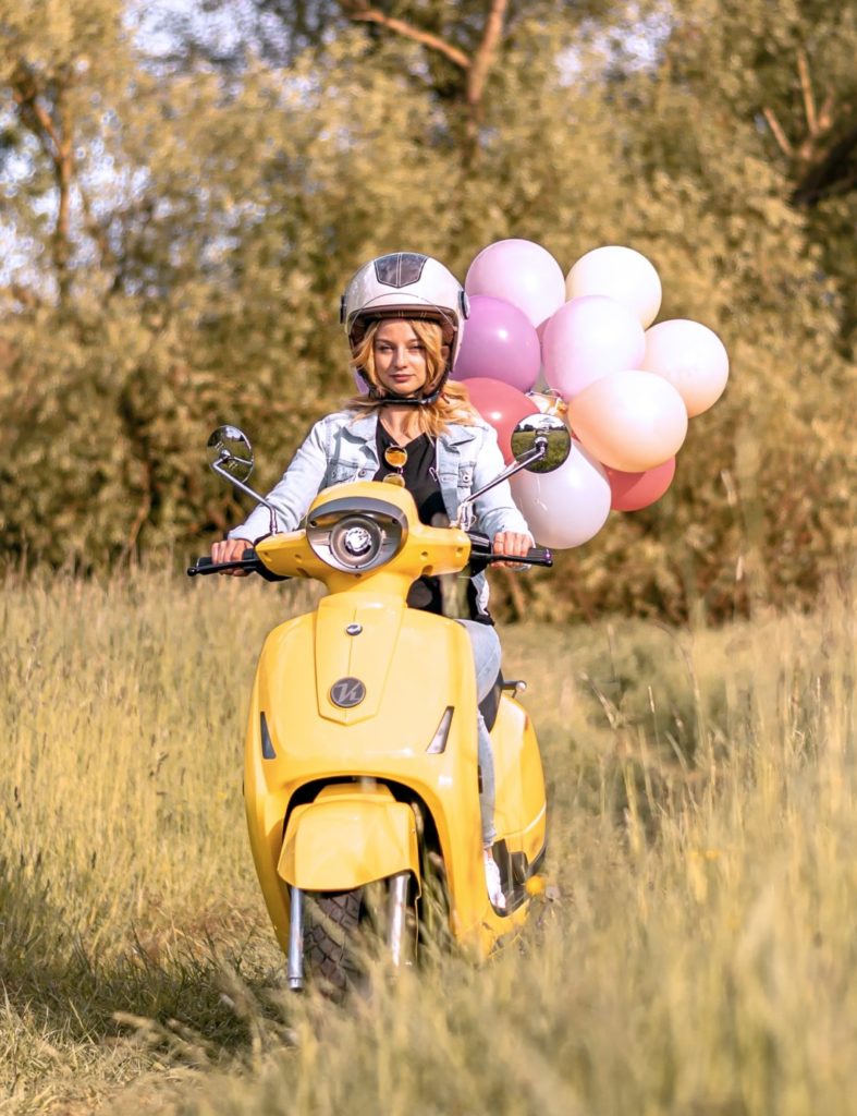 girl on a yellow scooter in a field wearing a white jacket and white open face helmet trailing pink balloons