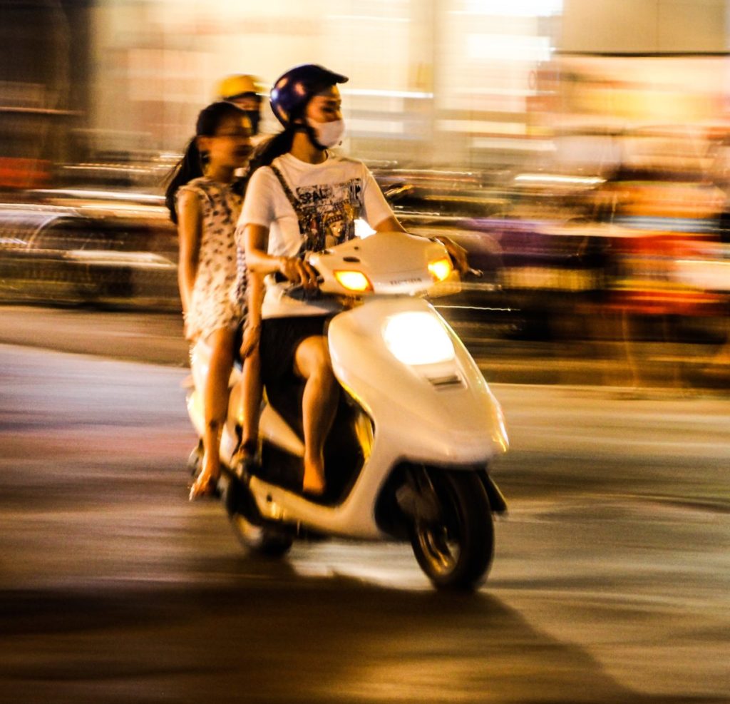 two black-haired women in white tee shirts and a child on a white scooter in a city at night